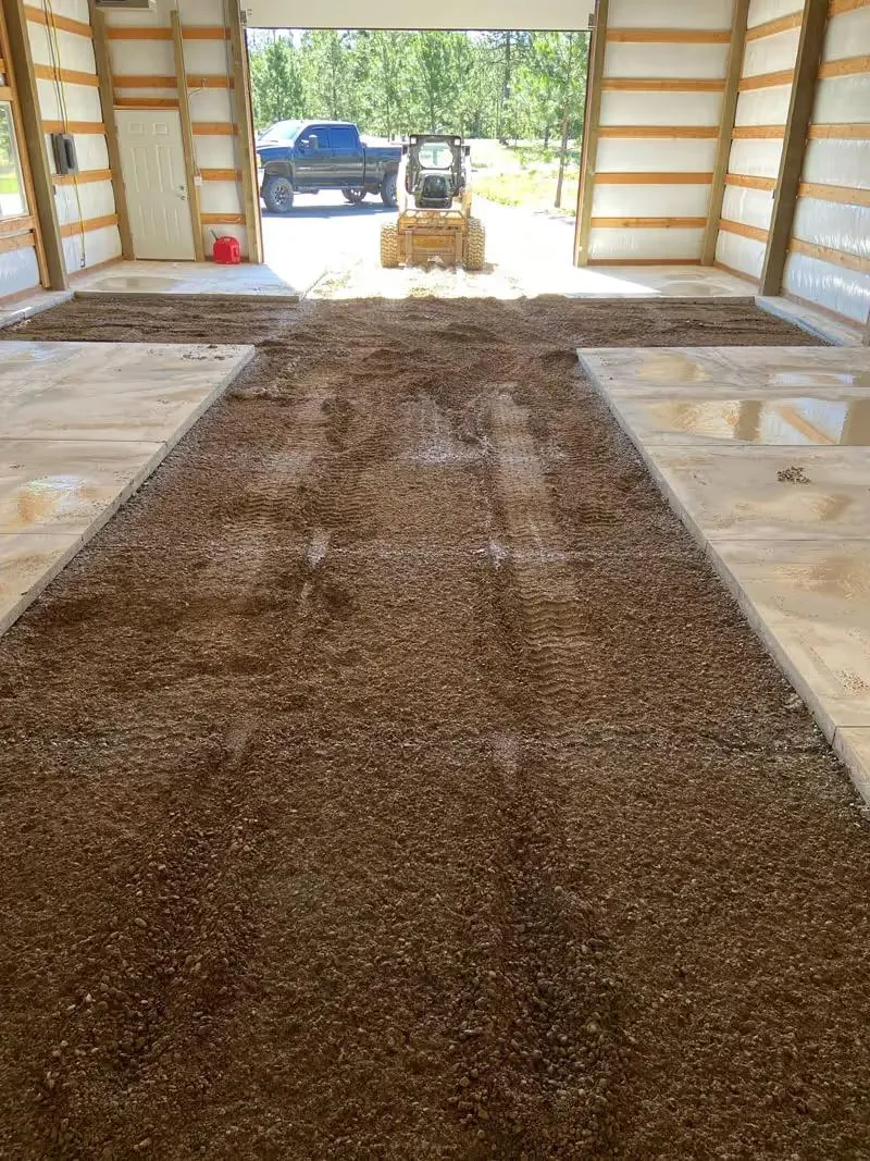 Gravel base spread across the floor of a pole building, looking out the open door to a skid steer