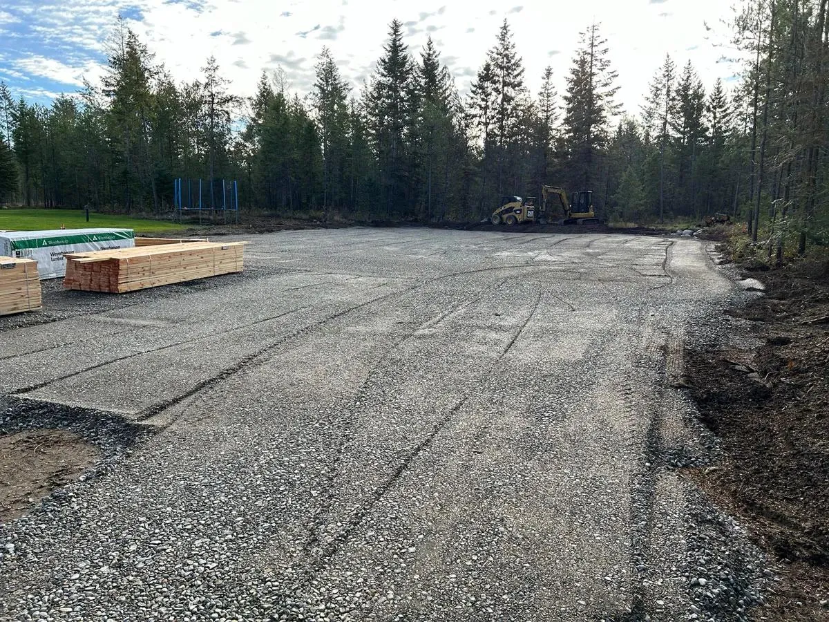 Freshly graded gravel pad surrounded by evergreen trees with an excavator in the background